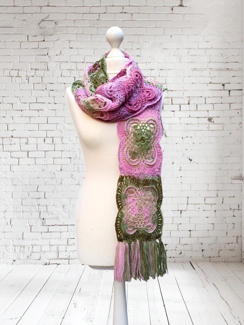 May include: A handmade crochet scarf in shades of pink, green, and olive. The scarf features a detailed floral pattern and a fringed edge. The scarf is displayed on a white mannequin against a white brick wall.