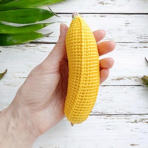 Puede incluir: Un juguete de pl&aacute;tano de crochet amarillo con un tallo rosa. El pl&aacute;tano se sostiene en una mano sobre un fondo de madera blanco.