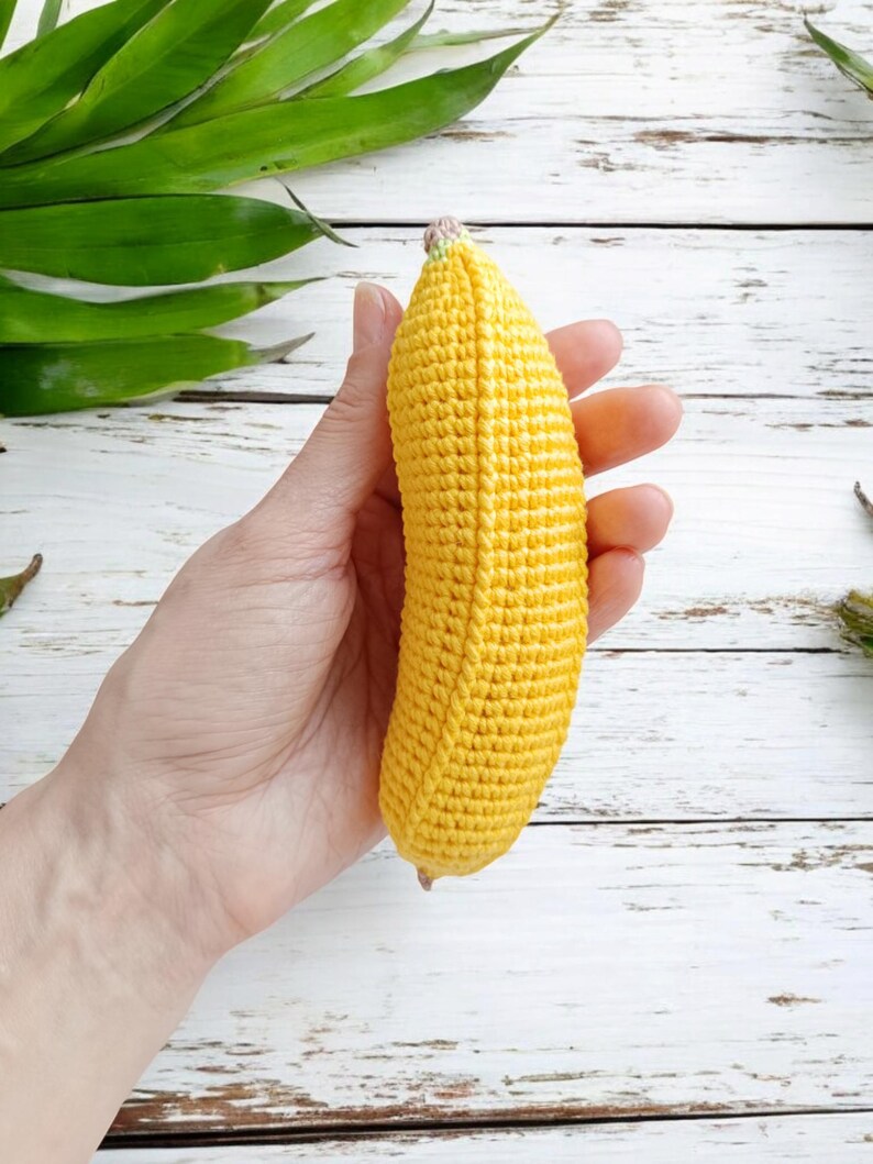 Puede incluir: Un juguete de pl&aacute;tano de crochet amarillo con un tallo rosa. El pl&aacute;tano se sostiene en una mano sobre un fondo de madera blanco.