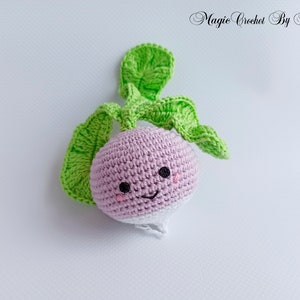 May include: A crocheted plush toy shaped like a turnip with a pink body, white bottom, and green leaves. The turnip has a smiling face with black eyes and pink cheeks.