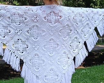 Puede incluir: Un chal blanco de ganchillo con un diseño calado detallado. El chal es triangular, con flecos en el borde inferior. El intrincado patrón presenta formas florales y geométricas, creando un aspecto delicado y elegante. El chal se muestra al aire libre.