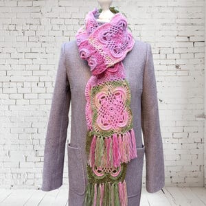 May include: A handmade, pink and green crochet scarf with a floral design and fringe. The scarf is draped over a gray coat on a mannequin. The scarf's colors include shades of pink, green, and a touch of yellow.