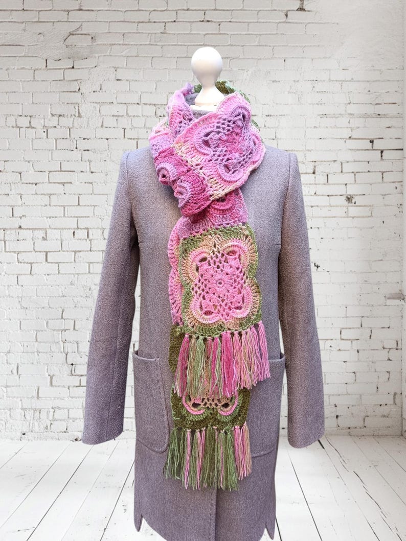 May include: A handmade, pink and green crochet scarf with a floral design and fringe. The scarf is draped over a gray coat on a mannequin. The scarf's colors include shades of pink, green, and a touch of yellow.