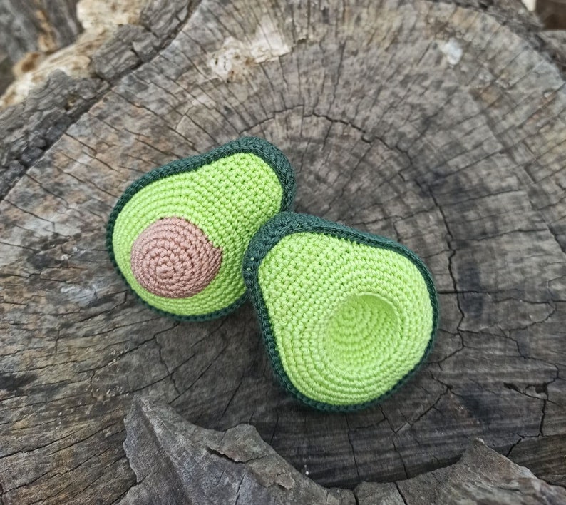 May include: Two crocheted avocado halves, one with a brown pit and the other with a green pit, on a wooden surface.