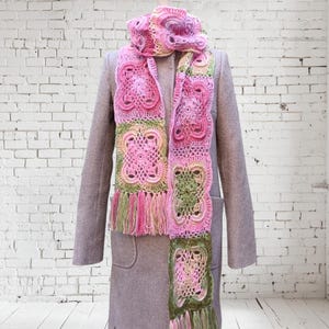 May include: A handmade crochet scarf in shades of pink, green, and cream, displayed on a mannequin. The scarf features a floral pattern and fringe. The mannequin is wearing a gray coat.
