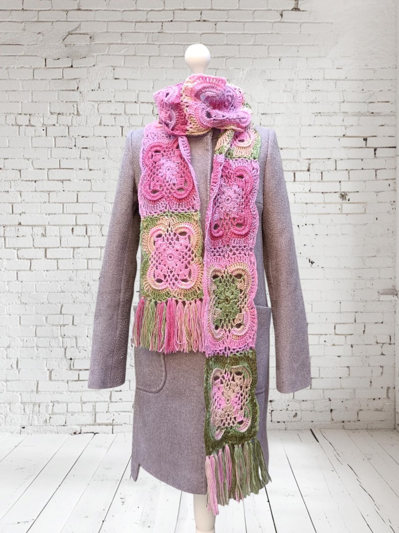 May include: A handmade crochet scarf in shades of pink, green, and cream, displayed on a mannequin. The scarf features a floral pattern and fringe. The mannequin is wearing a gray coat.