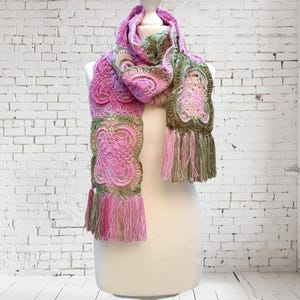 May include: A handmade crochet scarf in shades of pink and green, displayed on a mannequin. The scarf features intricate floral patterns and fringe detailing at the ends. The color palette includes various shades of pink and green.