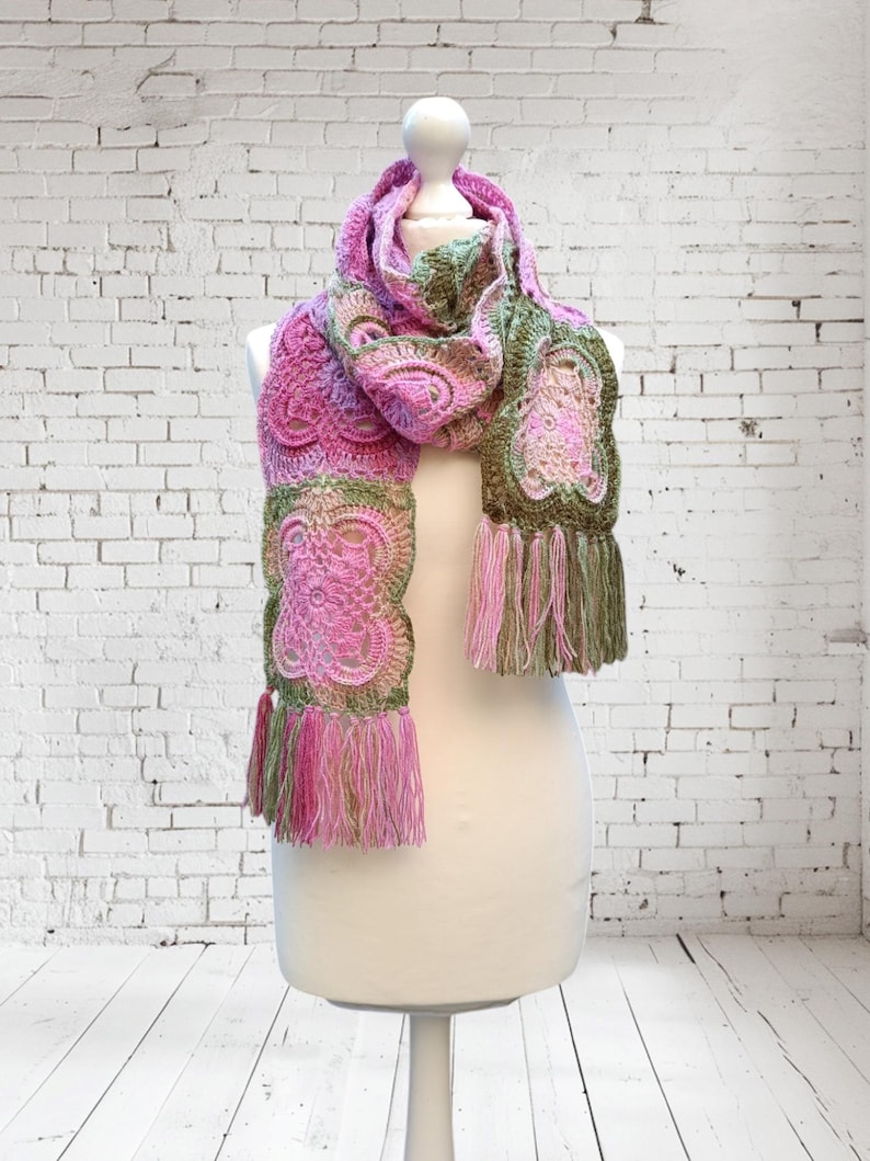May include: A handmade crochet scarf in shades of pink and green, displayed on a mannequin. The scarf features intricate floral patterns and fringe detailing at the ends. The color palette includes various shades of pink and green.
