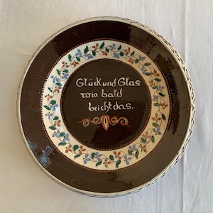 May include: A round, brown ceramic plate with a decorative floral border and the German text "Glück und Glas wie bald bricht das." The plate features a white inner ring with colorful flowers and a decorative edge.