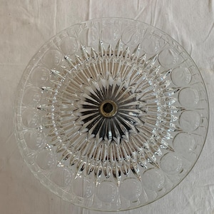 Op de afbeelding: Een glazen taartplateau van helder glas met een decoratief stervormig patroon en een metalen middenpaal. Het plateau is ontworpen om taarten, gebak of andere desserts te serveren.