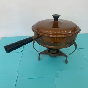 May include: A brown metal chafing dish with a black handle and a black knob on the lid. The dish is on a stand with three legs.