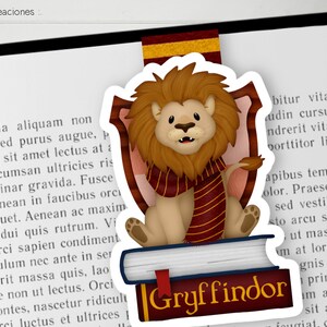 Op de afbeelding: Een Gryffindor-bladwijzer met een cartoon-leeuw die een rood-gouden sjaal draagt, zittend op een stapel boeken. De bladwijzer is wit met een rood-gouden rand en het woord "Gryffindor" in gouden letters.
