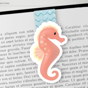 Può includere: Un segnalibro a forma di cavalluccio marino rosa e giallo con un contorno bianco. Il segnalibro è a forma di cavalluccio marino con una coda arricciata e una pinna sulla schiena. Il cavalluccio marino è su uno sfondo bianco con un motivo ondulato blu e bianco dietro.