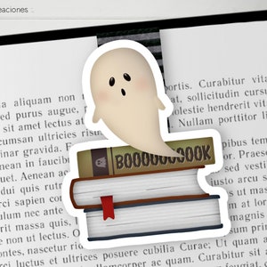 Può includere: Un adesivo di fantasma bianco con un'espressione sorpresa, che fluttua sopra una pila di tre libri. Il libro in cima ha la parola "BOO" scritta in marrone.