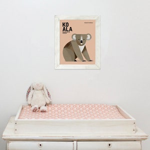 K&ouml;nnte beinhalten: Ein gerahmter Druck mit einer Koala-Illustration und dem Wort &bdquo;KOALA&ldquo; in fetten Buchstaben. Der Druck befindet sich in einem wei&szlig;en Rahmen und h&auml;ngt &uuml;ber einer wei&szlig;en Wickelkommode mit einer rosa gemusterten Wickelauflage. Ein Stoffhase sitzt auf der Wickelkommode.