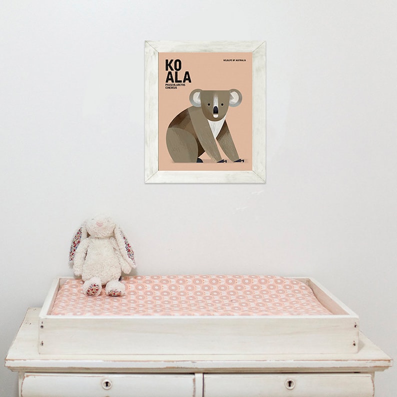 K&ouml;nnte beinhalten: Ein gerahmter Druck mit einer Koala-Illustration und dem Wort &bdquo;KOALA&ldquo; in fetten Buchstaben. Der Druck befindet sich in einem wei&szlig;en Rahmen und h&auml;ngt &uuml;ber einer wei&szlig;en Wickelkommode mit einer rosa gemusterten Wickelauflage. Ein Stoffhase sitzt auf der Wickelkommode.