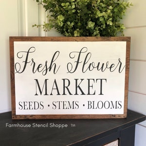 May include: A white wooden sign with a brown wood frame. The sign says "Fresh Flower Market" in black cursive lettering. Below the market name, it says "Seeds. Stems. Blooms" in black lettering.