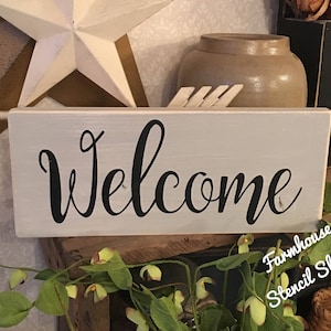 May include: White wooden sign with the word "Welcome" written in black cursive script. The sign is on a shelf with green plants and a brown wicker basket.