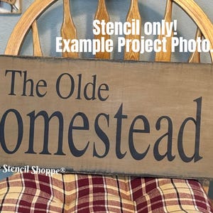 May include: A wooden sign with the words "The Olde Homestead" in dark blue lettering and a black star. The sign is a light brown color and has a distressed look. The text "Stencil only! Example Project Photo" is at the top.
