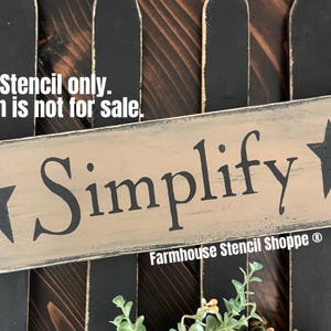 May include: A rustic wooden sign with the word "Simplify" painted in black lettering. Two black stars are on either side of the word. The sign is distressed and has a light brown background. The text "Stencil only. Sign is not for sale." is visible above the sign. The text "Farmhouse Stencil Shoppe®" is visible below the sign.