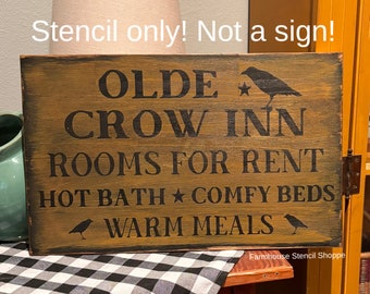 May include: A wooden sign with black painted text that reads "Olde Crow Inn Rooms for Rent Hot Bath Comfy Beds Warm Meals" with a crow silhouette on each side of the text and a star above the word "Bath". The sign has a distressed, rustic look and is labeled "Farmhouse Stencil Shoppe".