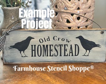 May include: A rectangular wooden sign with a distressed gray background features the words "Old Crow Homestead" in black lettering, flanked by black crow silhouettes. The sign is decorated with a black and white checkered ribbon.