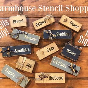 May include: A collection of wooden signs with winter-themed words and phrases, including "Snowman", "Snowflakes", "Cozy", "Sledding", "Peace", "Winter", "Let it Snow", "Frosty", "Joy", "Hot Cocoa", "Brrr!" and "Believe". The signs are decorated with burlap ribbon, twine, and small wooden stars. The signs are arranged on a wooden surface. The text "Stencils Only!" and "Not Signs!" are written on the image.