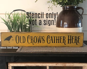 May include: A yellow wooden sign with black lettering that reads "Old Crows Gather Here". A black silhouette of a crow is to the left of the text. The sign is on a black surface with a white and black checkered fabric in front of it.