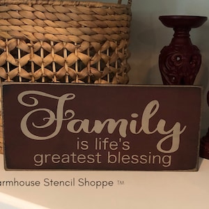 May include: A brown wooden sign with white text that reads "Family is life's greatest blessing". The sign is sitting on a shelf with a wicker basket and two red candle holders.