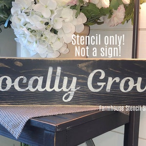 May include: A black wooden sign with white lettering that reads "Locally Grown". The sign is distressed and has a rustic look. The text "Stencil only! Not a sign!" is written in white above the main text. The sign is sitting on a black table with a white and gray checkered tablecloth.