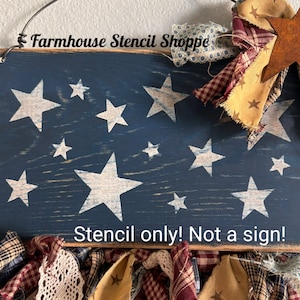 May include: A blue wooden sign with white stars painted on it. The sign has a distressed look and is decorated with fabric scraps. The text "Farmhouse Stencil Shoppe" and "Stencil only! Not a sign!" is printed on the sign.