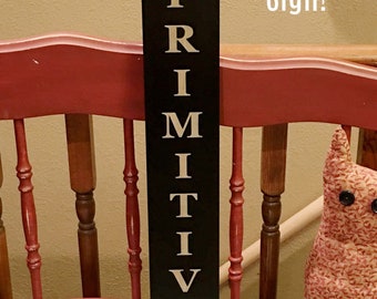 May include: Black wooden sign with gold lettering spelling "PRIMITIVES" with a gold star at the top and bottom. The sign is on a red chair with a floral patterned pillow. The text "Stencil Only! Not a Sign!" is written in white on a beige wall. The text "Farmhouse Stencil Shoppe" is written in white at the bottom of the sign.