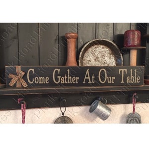 May include: A black wooden sign with the text "Come Gather At Our Table" in a light beige color. A decorative bow is attached to the left side. The sign is part of a shelf display with other rustic kitchen items.