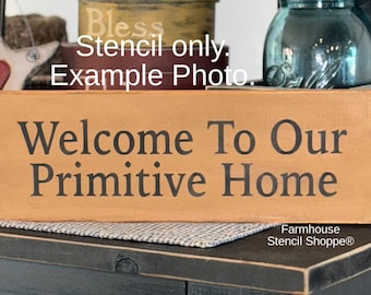 May include: A rectangular wooden sign with the text "Welcome To Our Primitive Home" in black lettering. The sign is light brown and sits on a dark surface. The words "Stencil only. Example Photo." are above the sign.