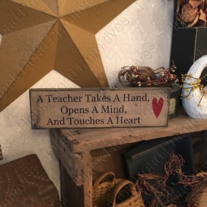 May include: A wooden sign with a red heart painted on it reads "A Teacher Takes A Hand, Opens A Mind, And Touches A Heart".