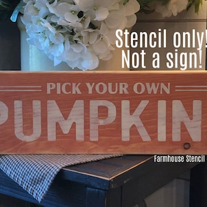 May include: A wooden sign with the text "PICK YOUR OWN PUMPKINS" in white letters on a red background. The sign is on a table with a checkered cloth. The text "Stencil only! Not a sign!" is written above the sign. The text "Farmhouse Stencil Shoppe" is written below the sign.