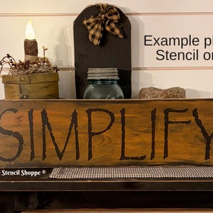 May include: A wooden sign with the word "SIMPLIFY" painted in black lettering on a brown background. The sign is sitting on a table with other decorative items.
