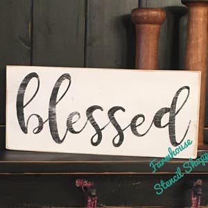 May include: White wooden sign with the word "blessed" written in black script. The sign is on a black shelf with a wooden dowel behind it.