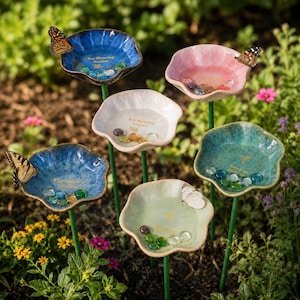 May include: A collection of six ceramic flower-shaped garden stakes in blue, pink, white, and green, each featuring a central basin filled with small glass pebbles and gold text, supported by thin green metal stems, with butterflies resting on rims.