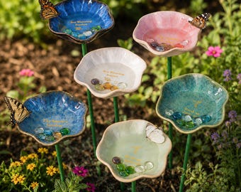 Könnte beinhalten: Ein Set aus sechs blumenförmigen Gartensteckern aus Keramik in Blau, Rosa, Weiß und Grün, mit einer zentralen Schale, die mit kleinen Glassteinen und goldenem Text gefüllt ist, auf dünnen grünen Metallstäben, mit Schmetterlingen an den Rändern.