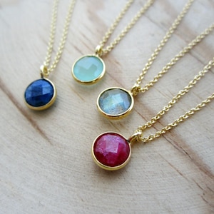 May include: Three gold necklaces with different colored gemstone pendants. The pendants are round and have a faceted design. The necklaces are on a wooden surface.
