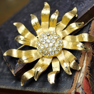 May include: A gold-toned brooch in the shape of a flower with a center made of clear rhinestones.