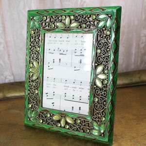 May include: A decorative green picture frame with ornate detailing and embedded green gemstones. The frame surrounds sheet music with handwritten notes. The frame's design includes diamond and leaf patterns, with a metallic filigree border.