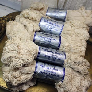 May include: Several skeins of off-white crochet and knitting cotton are stacked on a decorative tray. Each skein is wrapped with a silver and blue label that reads "Wondersheen Mercerized Crochet & Knitting Cotton". The cotton appears soft and ready for crafting.
