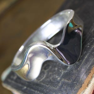 May include: A polished silver ring featuring a continuous, fluid wave-like design that curves along the band. The metal surface is highly reflective, creating smooth, undulating contours that wrap around the finger in a singular, flowing shape.
