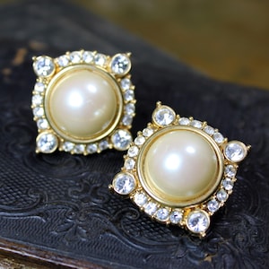 以下が含まれることがあります： 中央に大きな丸いクリーム色の真珠をあしらった、正方形の金色のイヤリング2つ。真珠は透明で輝くクリスタルのリングで囲まれています。イヤリングは、暗く質感のある表面に置かれています。