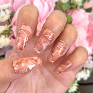 May include: Close-up of a hand wearing a set of peach-colored press-on nails with a white marble design.