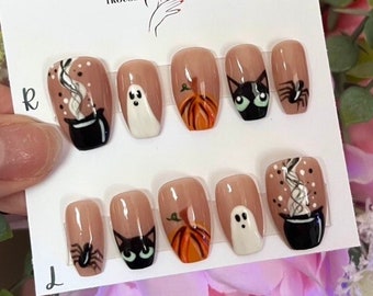 Puede incluir: Un conjunto de diez uñas postizas con temática de Halloween. Las uñas son de color nude y presentan varios diseños, incluyendo un caldero negro con vapor, un fantasma blanco, una calabaza naranja, un gato negro y una araña. Las uñas están dispuestas en dos filas de cinco, con el texto "Lady Trousse" visible en la parte superior de la tarjeta.