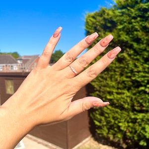 May include: A hand with long, light pink nails with a marbled design. The nails are long and almond shaped. The hand is wearing a thin silver ring with small clear stones.