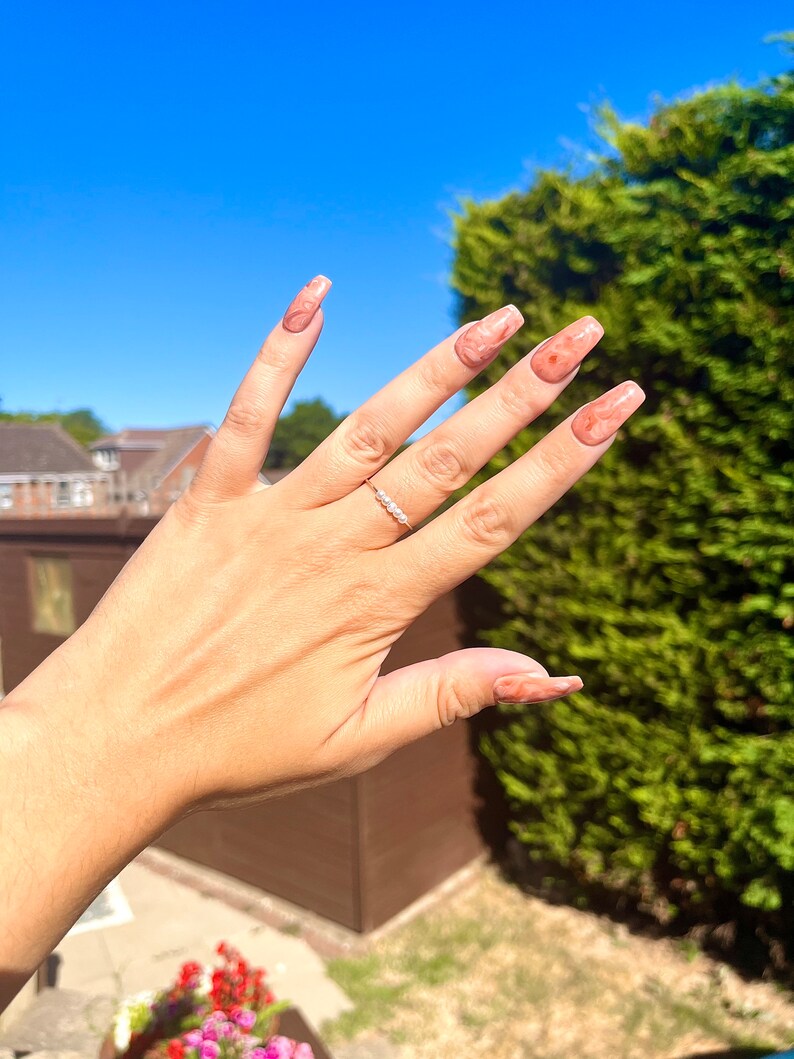 May include: A hand with long, light pink nails with a marbled design. The nails are long and almond shaped. The hand is wearing a thin silver ring with small clear stones.